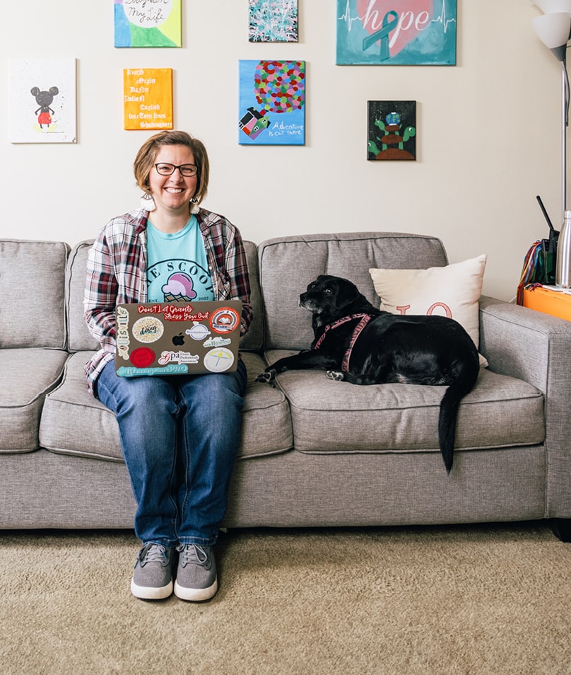 Grant writing professional Bethany Planton sits on a gray couch in front of a wall hung with bright, colorful pictures. She is smiling and wearing blue jeans. She has a laptop on her lap and a black dog with a silver snout next to her.