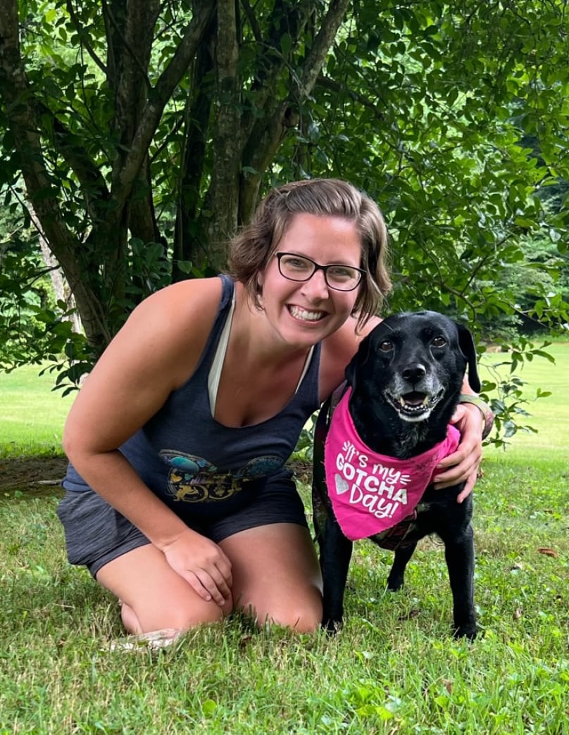 Grant writing professional Bethany Planton kneels in the grass next to a black dog with a silver snout. The dog wears a hot pink bandana that reads "It's my gotcha day!" Both are smiling.
