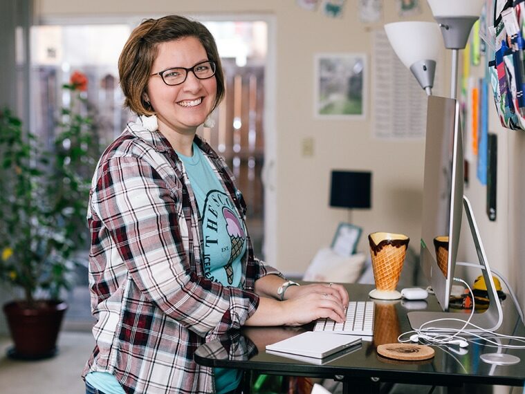 Bethany Planton, expert in grant writing for nonprofits, is pictured from the side as she works on a computer at a standing desk. She is smiling and wearing a flannel shirt over a graphic tee with an ice cream cone on it.