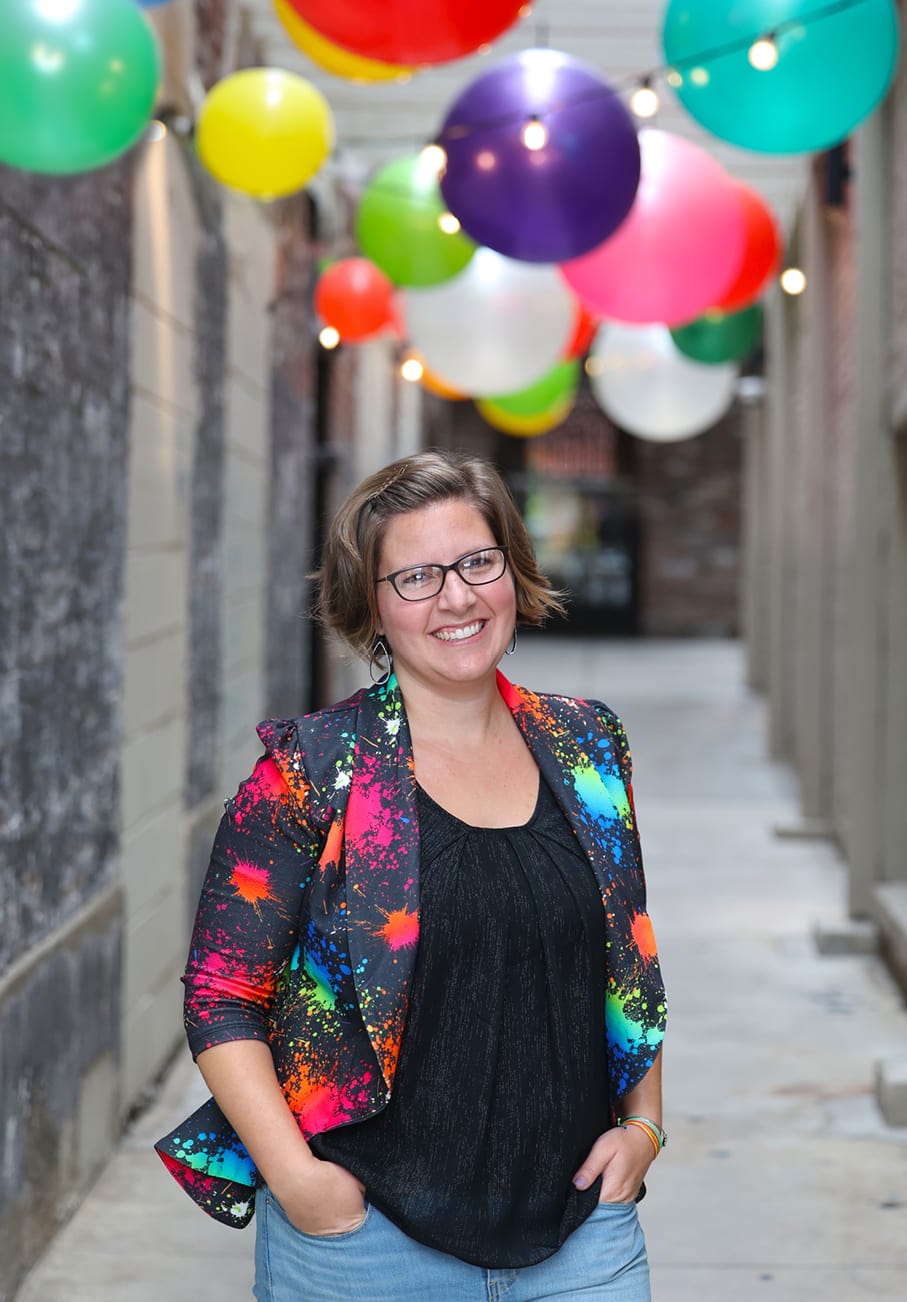 Bethany Planton, expert in grant writing for nonprofits, smiles while standing in a concrete alleyway with colorful round string lights strung above.