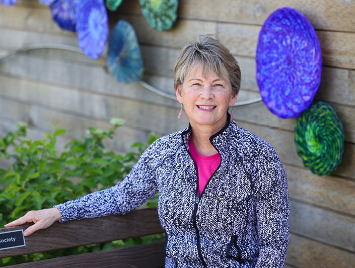 Grant writing professional Melissa Draut smiles while sitting on a wooden bench in front of an exterior wall adorned with brightly colored circular art pieces.