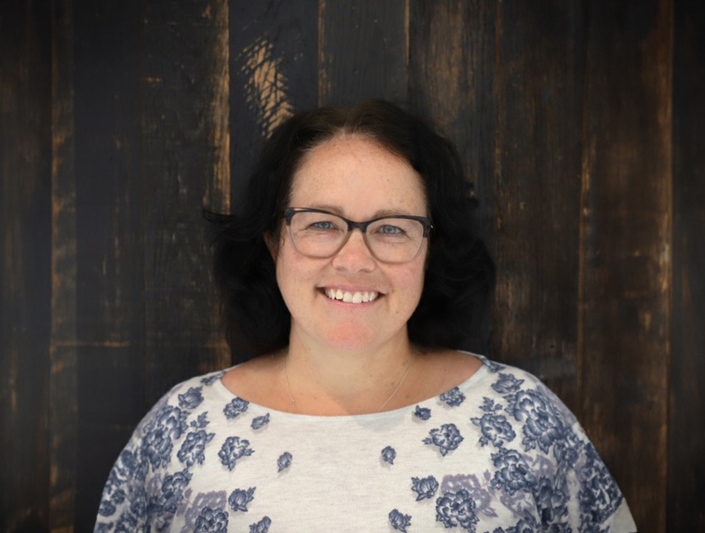 Grant writing professional Vickie Johnson smiles in front of a rustic wooden wall. She has glasses, shoulder length dark hair, and a white blouse with blue flowers on the sleeves.
